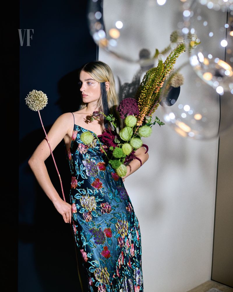 A blonde woman poses in a floral dress, holding flowers in front of a gray and black backdrop with illuminated translucent orbs.