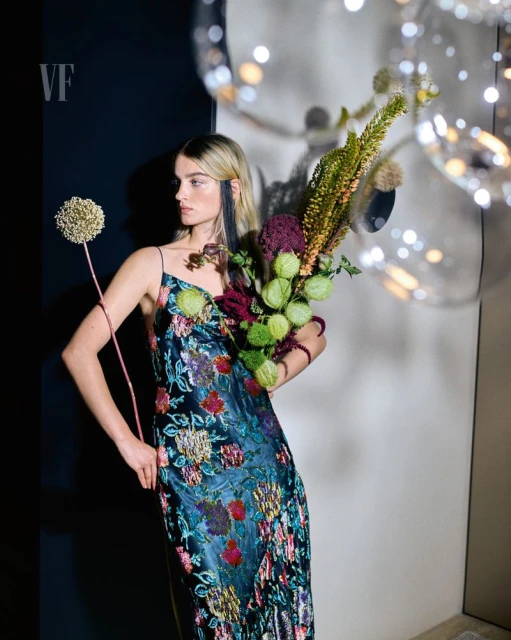 A blonde woman poses in a floral dress, holding flowers in front of a gray and black backdrop with illuminated translucent orbs.
