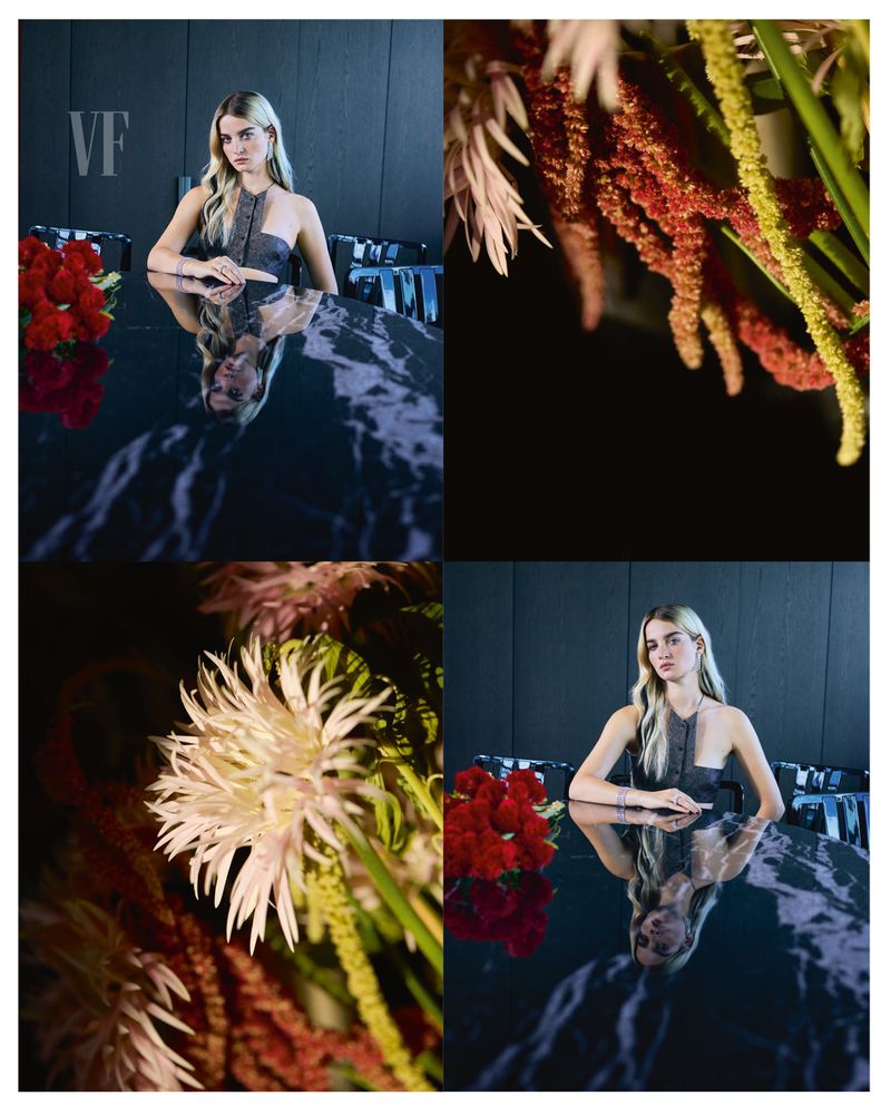 Blonde woman poses at a reflective black marble table with red roses and assorted greenery, illuminated by studio lighting.