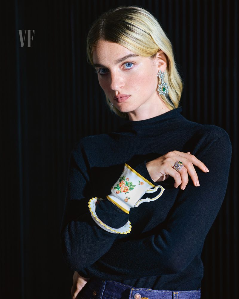 Elegant woman in a black turtleneck, adorned with floral teacup and diamond jewelry, poses against a dark backdrop for Vanity Fair.