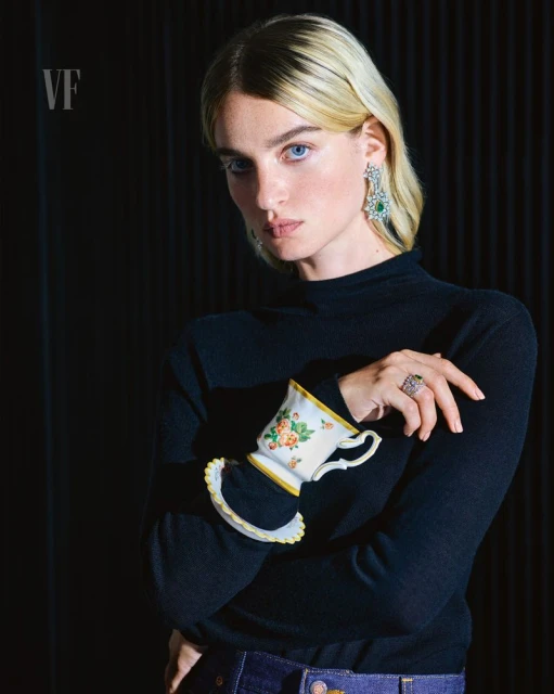 Elegant woman in a black turtleneck, adorned with floral teacup and diamond jewelry, poses against a dark backdrop for Vanity Fair.