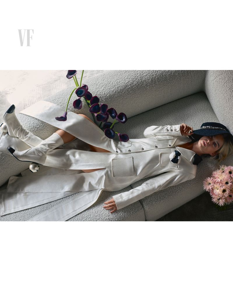 Fashion editorial portrait: Woman in white suit reclines on textured couch with black calla lilies, pink flowers, wearing a black hat.