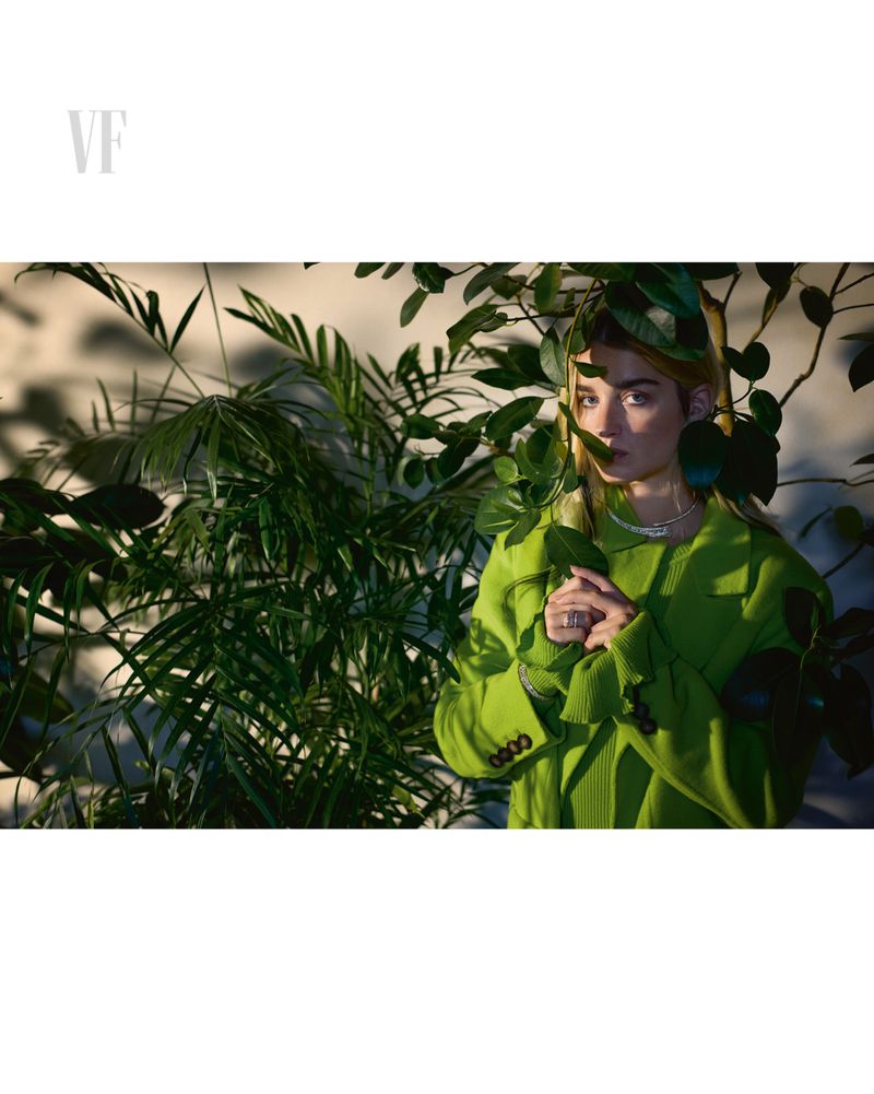 Stylish adult woman poses in a green outfit amongst lush plants inside a photo studio setting with soft, diffused natural lighting.