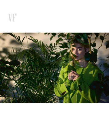 Stylish adult woman poses in a green outfit amongst lush plants inside a photo studio setting with soft, diffused natural lighting.