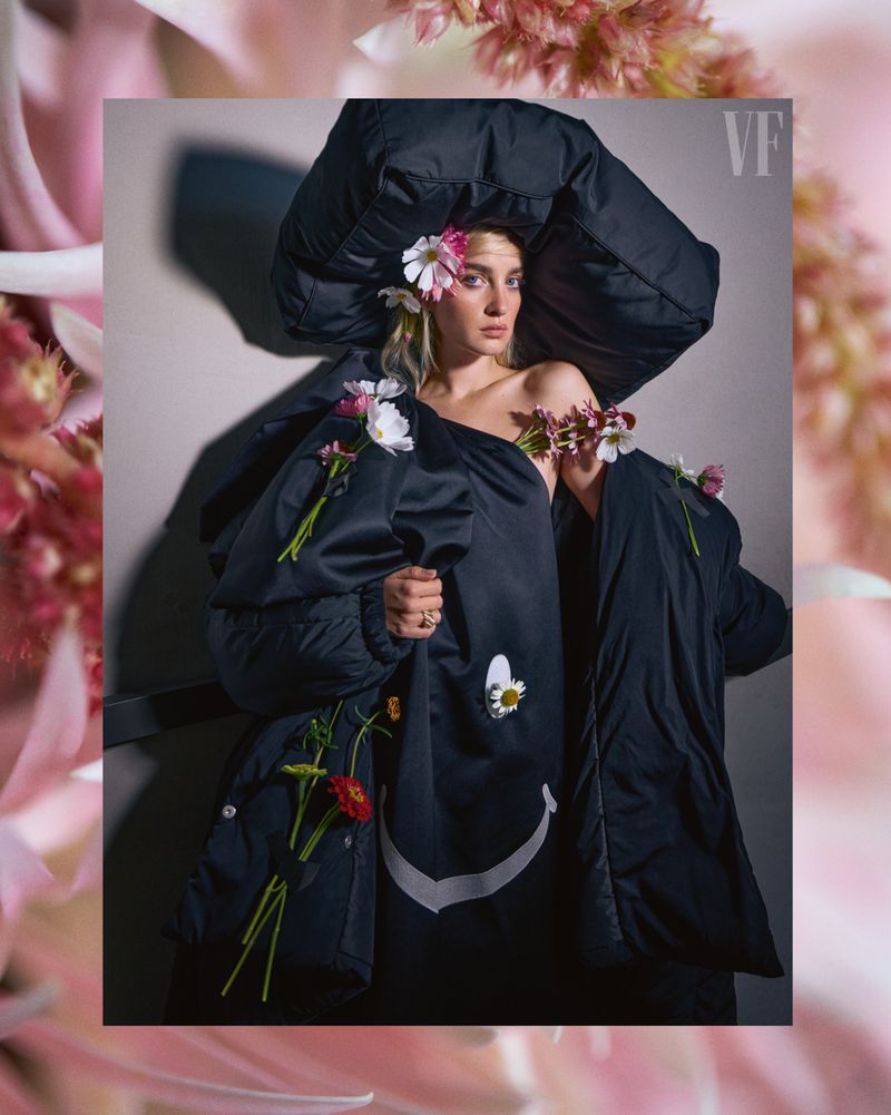 Woman poses in black coat with floral accents; studio setting. Fashion-forward with a blend of organic and urban elements, indoor portrait.
