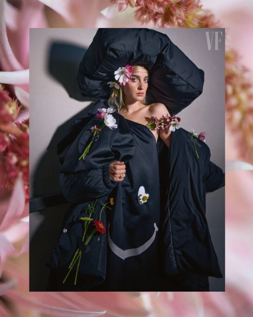 Woman poses in black coat with floral accents; studio setting. Fashion-forward with a blend of organic and urban elements, indoor portrait.