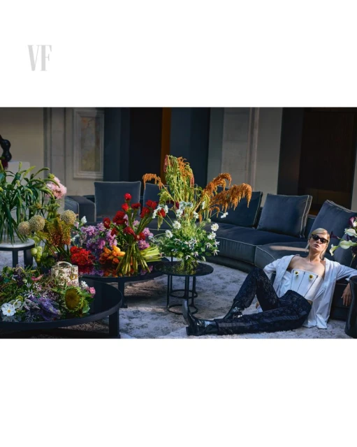 Indoor fashion shot of a woman reclining on a rug wearing a white top and black pants, surrounded by colorful flower arrangements.
