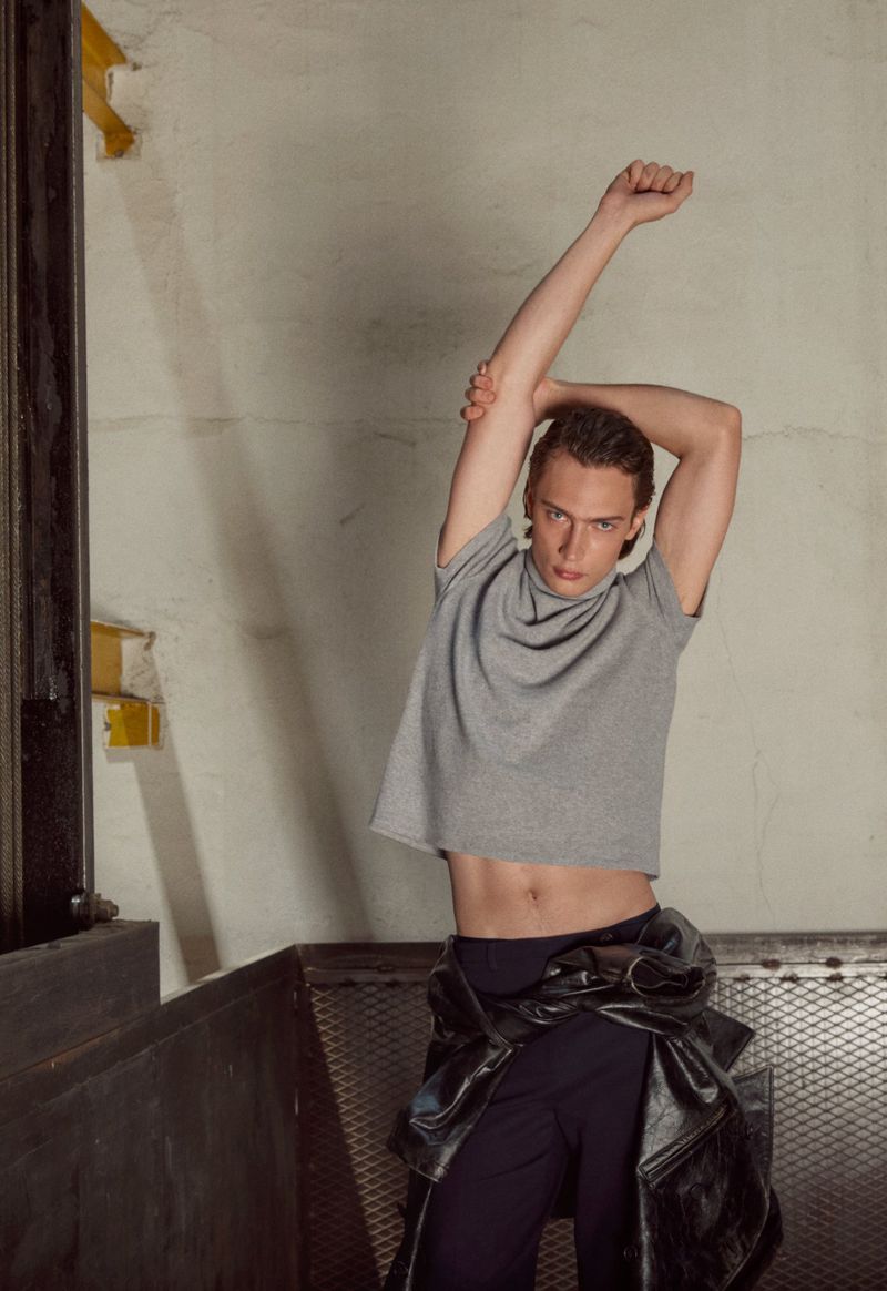 Young adult male stretches his arms above his head, wearing a grey t-shirt and black pants, set in an industrial interior.