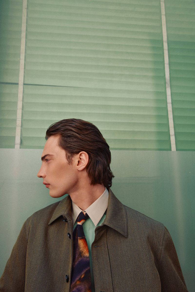 Fashion portrait of an adult male in a green-toned studio. He wears a gray coat and patterned tie, looking off to the side.