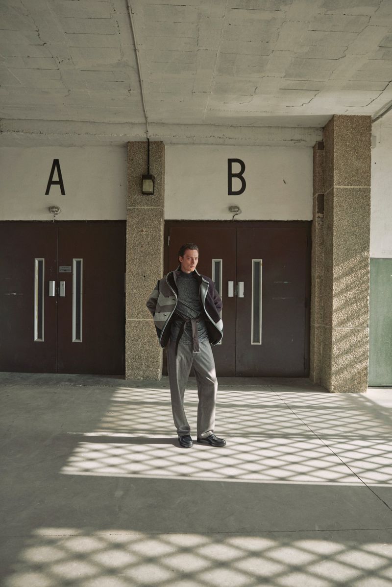 A man stands in front of doors marked A and B. He wears a jacket, sweater, trousers, and dress shoes in a building hallway.