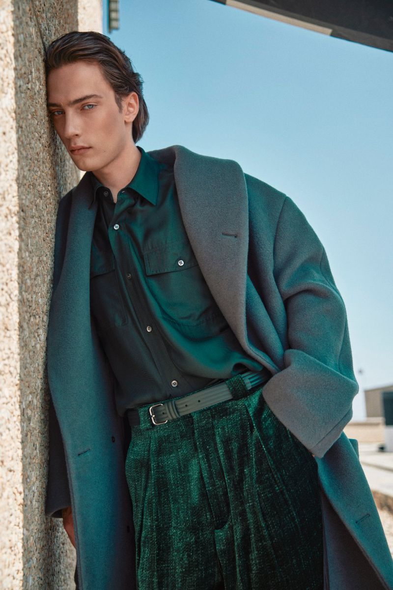Man in teal overcoat and shirt leans against wall in outdoor fashion shot, showcasing cool-toned fashion against a neutral backdrop under bright sky.