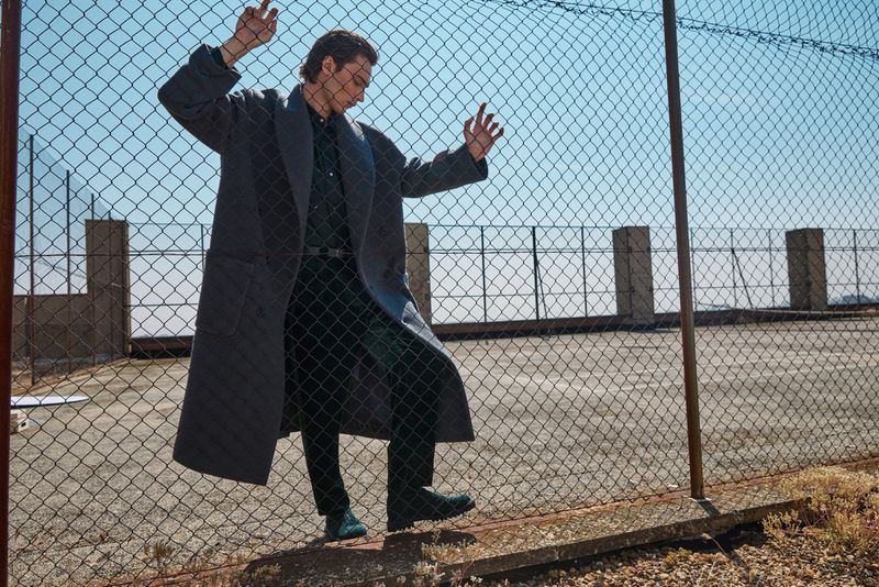 Model Freek Iven poses behind a chain-link fence wearing an oversized dark grey overcoat and black trousers under direct sunlight.