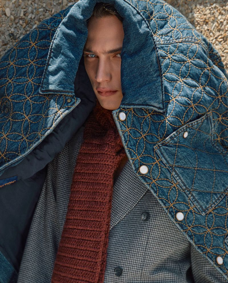 Close-up studio shot of a male model wearing a denim quilted jacket with a houndstooth wool jacket and a knit scarf, posing fashionably.