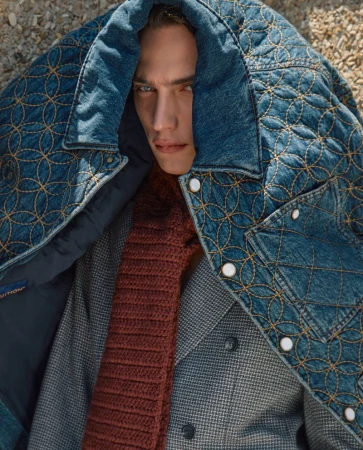 Close-up studio shot of a male model wearing a denim quilted jacket with a houndstooth wool jacket and a knit scarf, posing fashionably.