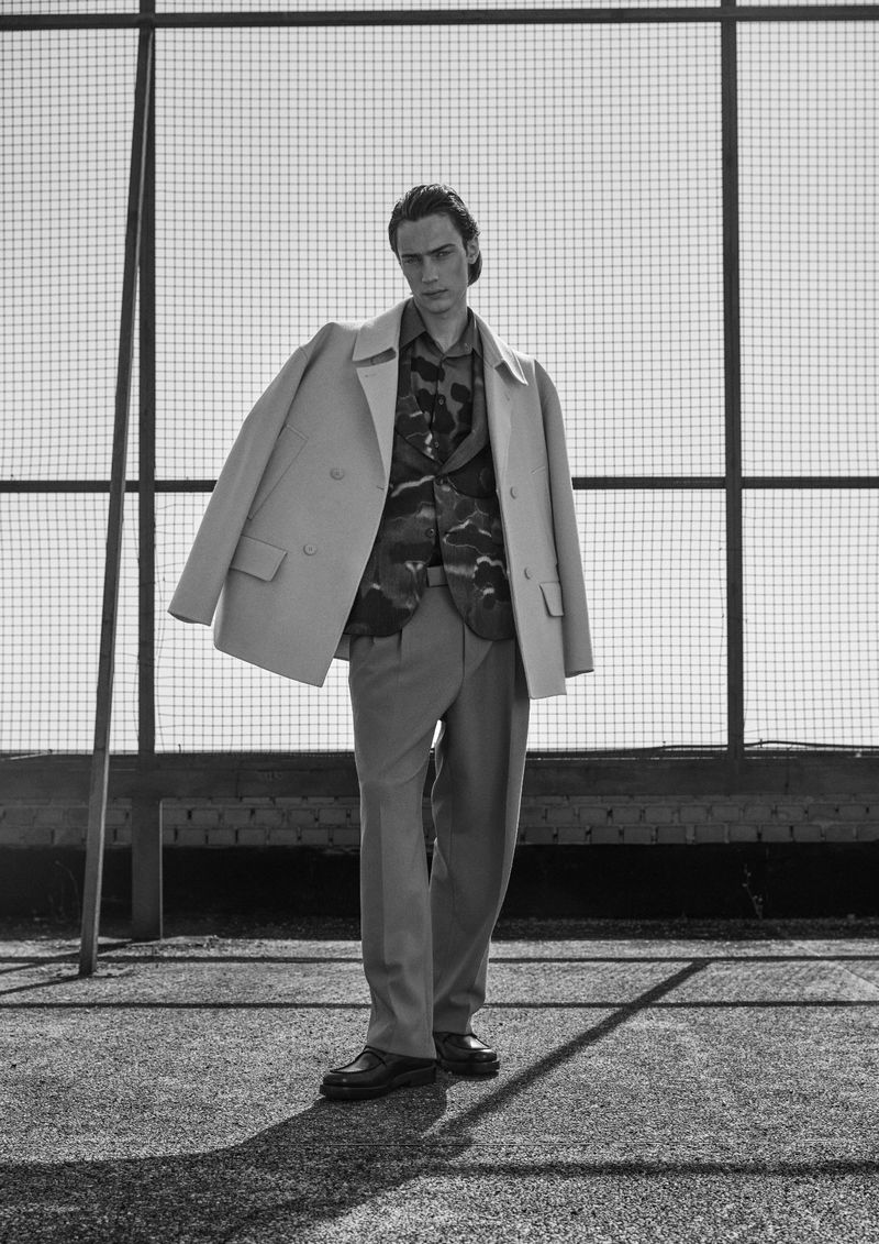 Model Freek Iven posing in a draped double-breasted overcoat, patterned jacket, and tailored trousers in front of a wire fence.