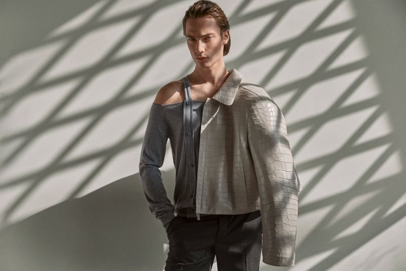 An adult male poses in a studio. He is wearing a gray top, black pants, and a leather jacket over his shoulder in a studio setting.