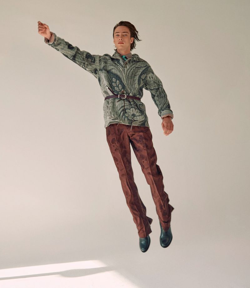 Fashionable young man in patterned jacket and pants leaps in a studio shot, highlighted by soft lighting and casting shadows.