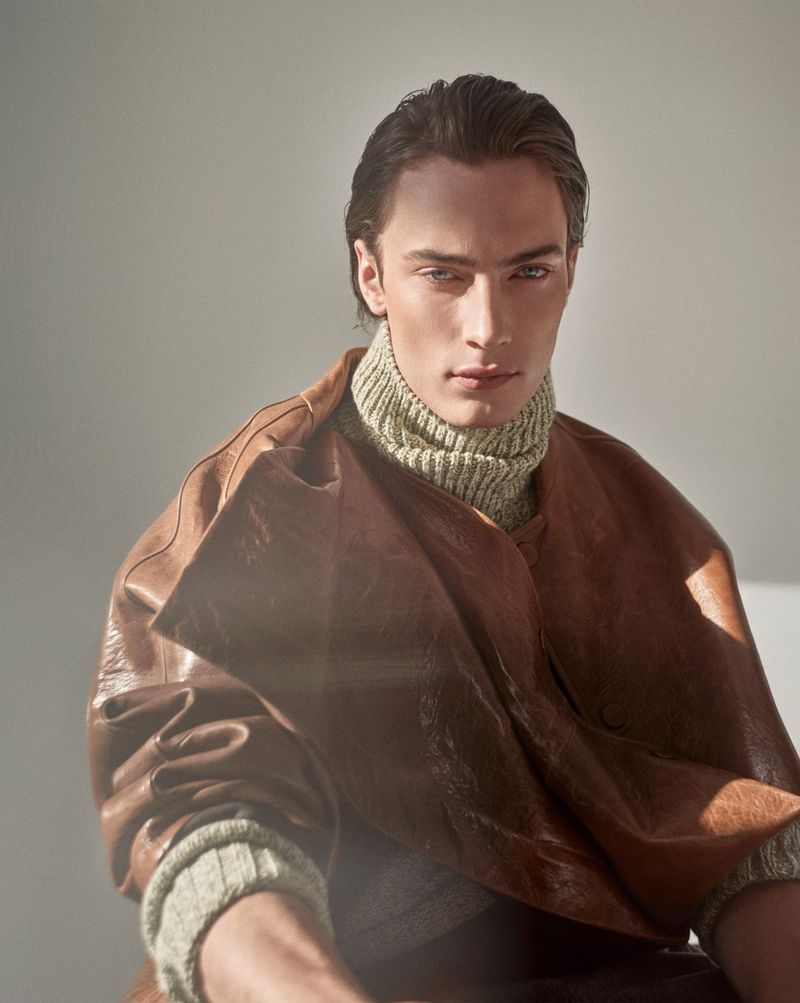 Portrait of a fair-skinned adult man with dark hair, wearing a brown leather jacket and ribbed wool sweater indoors with natural lighting.