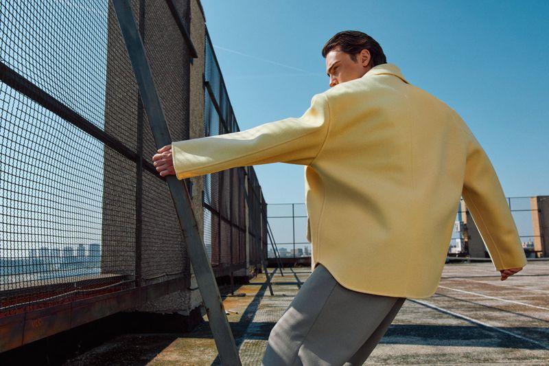 Man in yellow jacket on rooftop leans against metal fence with city buildings in distance against a blue sky in an eye-level shot.
