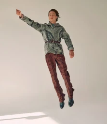 Model jumping mid-air wearing a patterned green jacket and brown trousers against a white studio background.