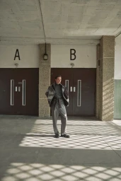 Model Freek Iven standing in a grey patterned jacket and trousers between two brown doors marked A and B with grid shadows on the floor.