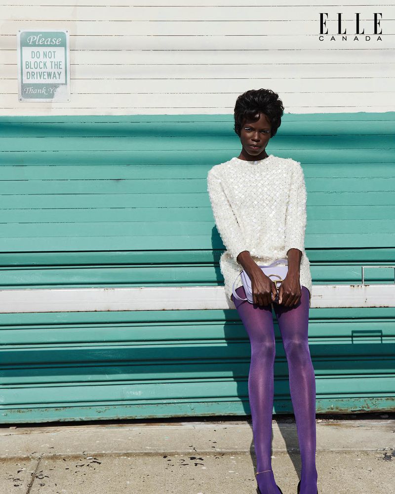 Fashion editorial shot of a Black female model in a white dress, purple tights, and heeled shoes against a teal garage door. Elle Canada logo visible.