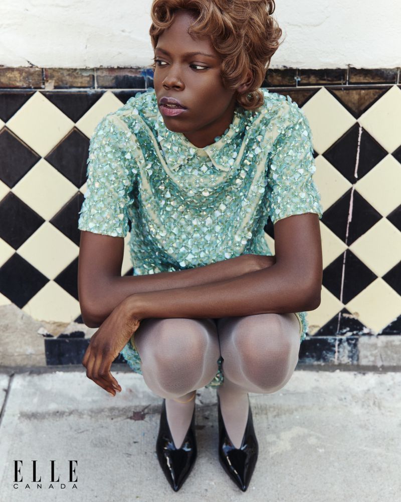 Editorial fashion shot of a Black woman in a sequin dress crouching against a tiled wall, glossy patent leather pumps, blonde retro hairstyle.