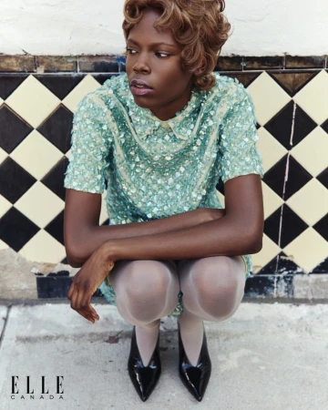 Editorial fashion shot of a Black woman in a sequin dress crouching against a tiled wall, glossy patent leather pumps, blonde retro hairstyle.