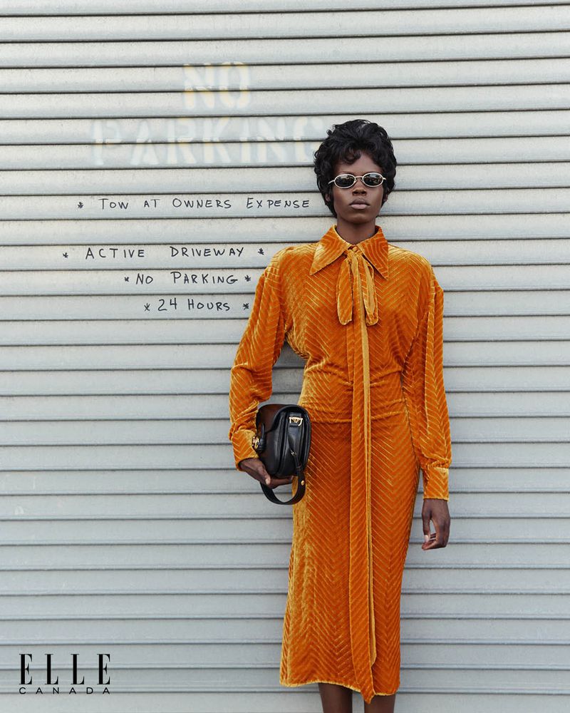 A woman poses in an orange skirt suit, holding a black purse in front of a metal shutter door with 'No Parking' signs.