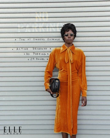 A woman poses in an orange skirt suit, holding a black purse in front of a metal shutter door with 'No Parking' signs.