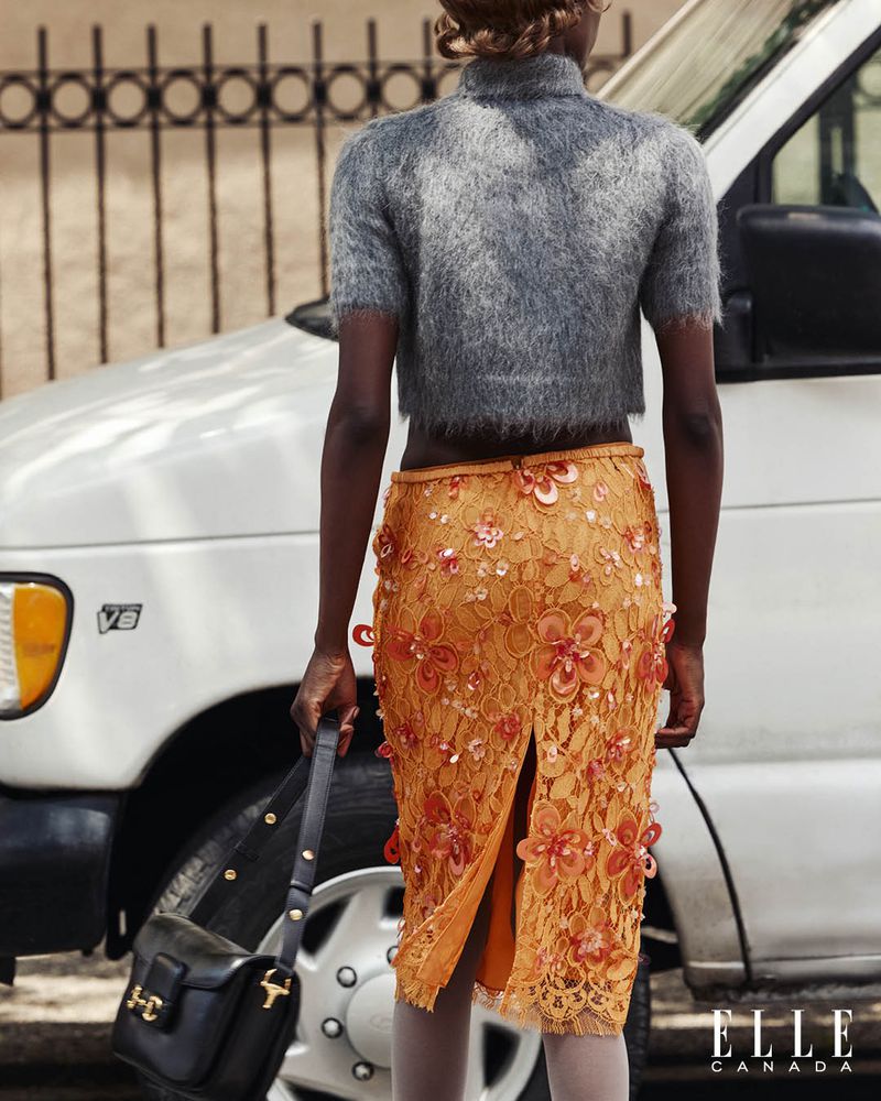 Woman from the rear wearing a grey cropped sweater and orange floral-applique skirt, holding a black purse, standing next to a white van.