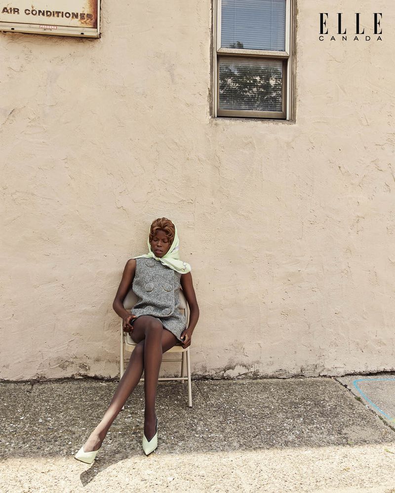 Elegant woman in a gray dress and headscarf sits on a chair outdoors against a textured wall, captured in a fashion editorial shot.