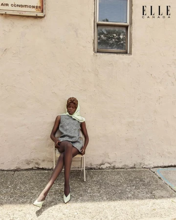 Elegant woman in a gray dress and headscarf sits on a chair outdoors against a textured wall, captured in a fashion editorial shot.