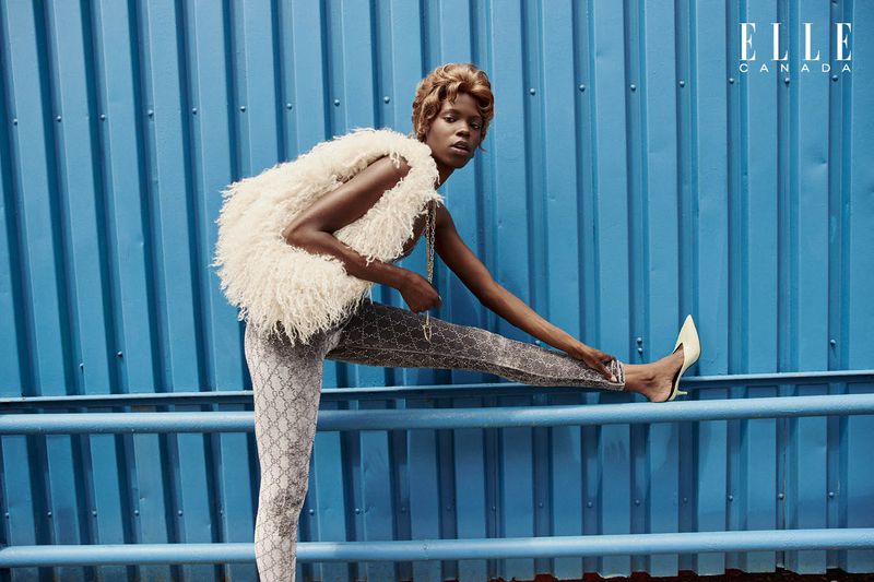 Fashion editorial photo: African-descent adult female model in a fur vest, patterned leggings, and heels against a blue background.