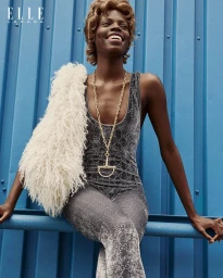 Agel Akol smiling in a grey patterned jumpsuit and white fur scarf against a blue corrugated wall for Elle Canada.