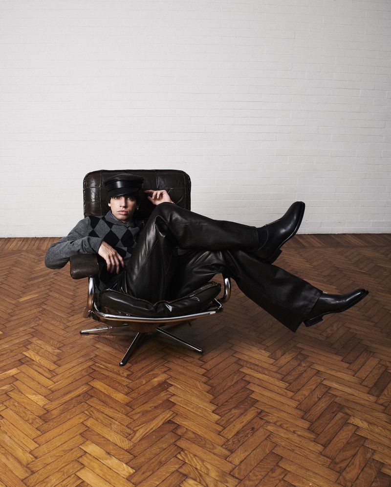 Young adult poses in leather pants and hat, seated on a modern chair in a minimalist interior setting, showcasing fashion-forward style.