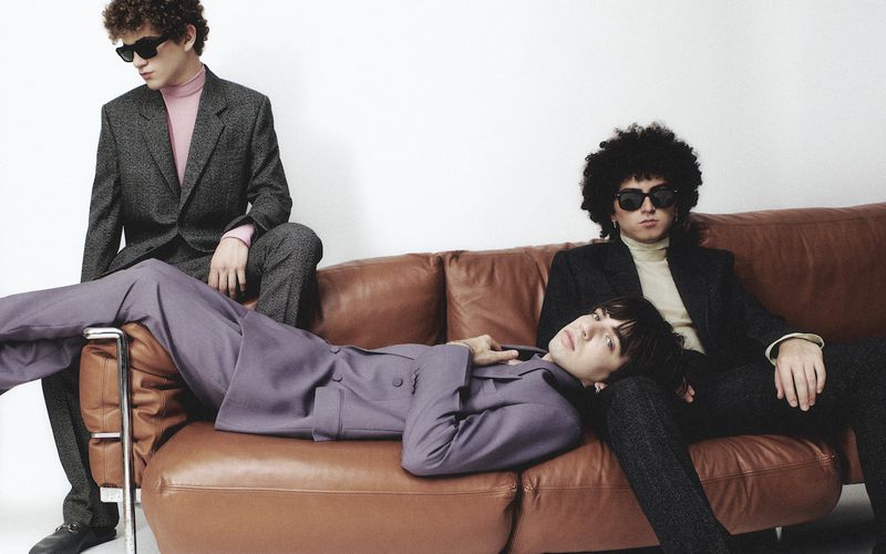 Three young adult men are posing on a brown leather couch indoors, wearing suits and sunglasses against a stark white background.
