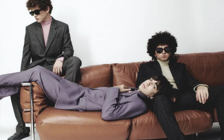Three young adult men are posing on a brown leather couch indoors, wearing suits and sunglasses against a stark white background.