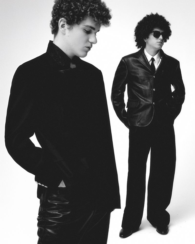 Two young men in dark suits stand in a photo studio, one with sunglasses, exuding an air of understated cool against a stark white backdrop.