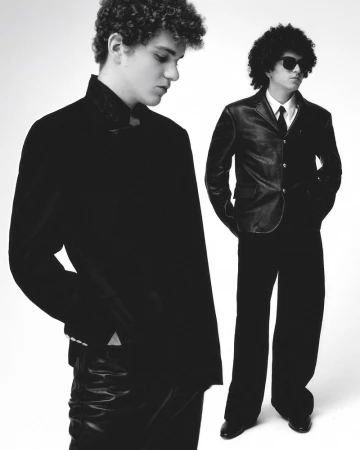 Two young men in dark suits stand in a photo studio, one with sunglasses, exuding an air of understated cool against a stark white backdrop.
