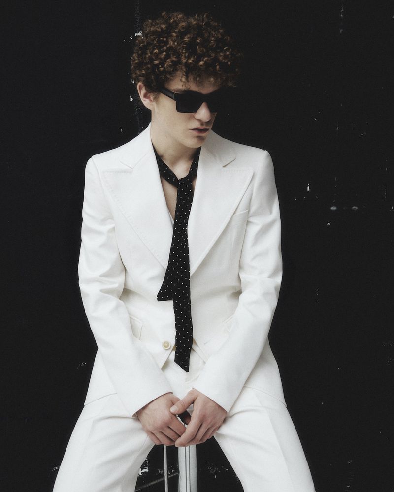Man with curly hair wearing a white suit and sunglasses, posing in a photo studio against a black background with natural window light.