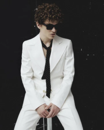 Man with curly hair wearing a white suit and sunglasses, posing in a photo studio against a black background with natural window light.