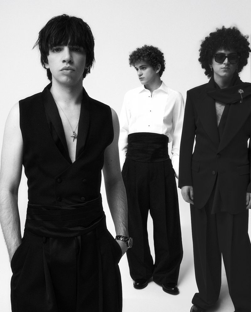 Three young adult males in formal black attire and fashionable hairstyles pose in a photo studio against a white backdrop.