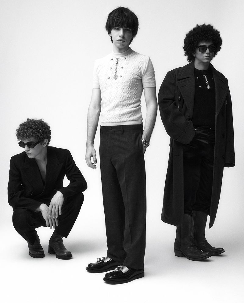 Black and white studio shot of three young adult male models wearing sunglasses, dark pants, coats and shiny shoes.