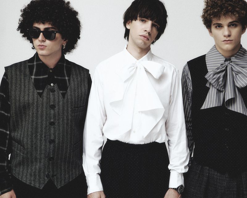 Three young adult males in formal attire, shirts with bows, and vests stand in front of a plain white background. Fashion studio portrait.