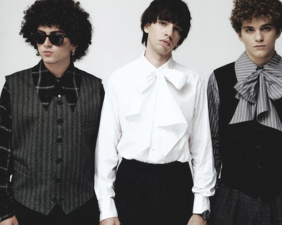 Three young adult males in formal attire, shirts with bows, and vests stand in front of a plain white background. Fashion studio portrait.