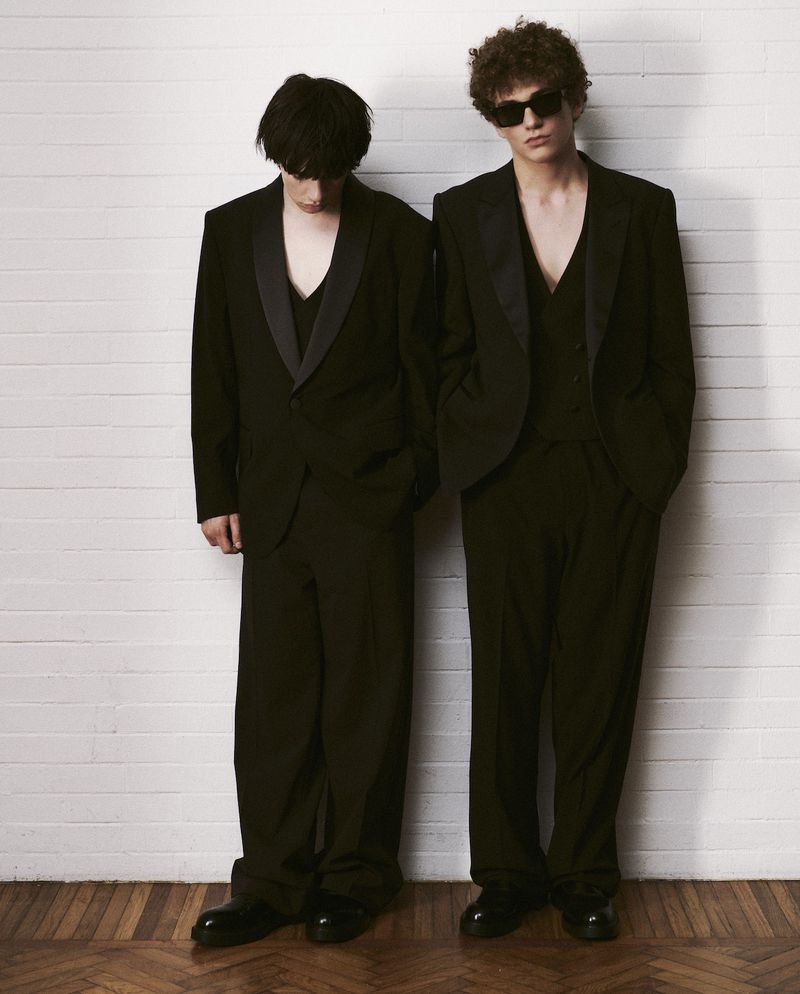 Two adult males in black suits stand against a white brick wall with a parquet wood floor, creating a high-contrast fashion editorial.