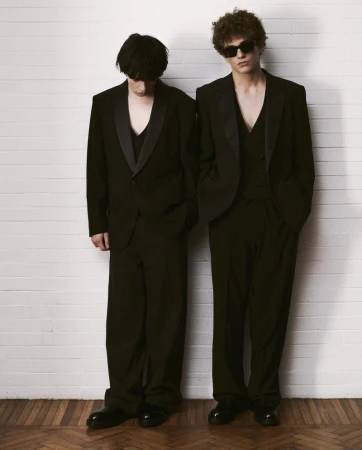 Two adult males in black suits stand against a white brick wall with a parquet wood floor, creating a high-contrast fashion editorial.
