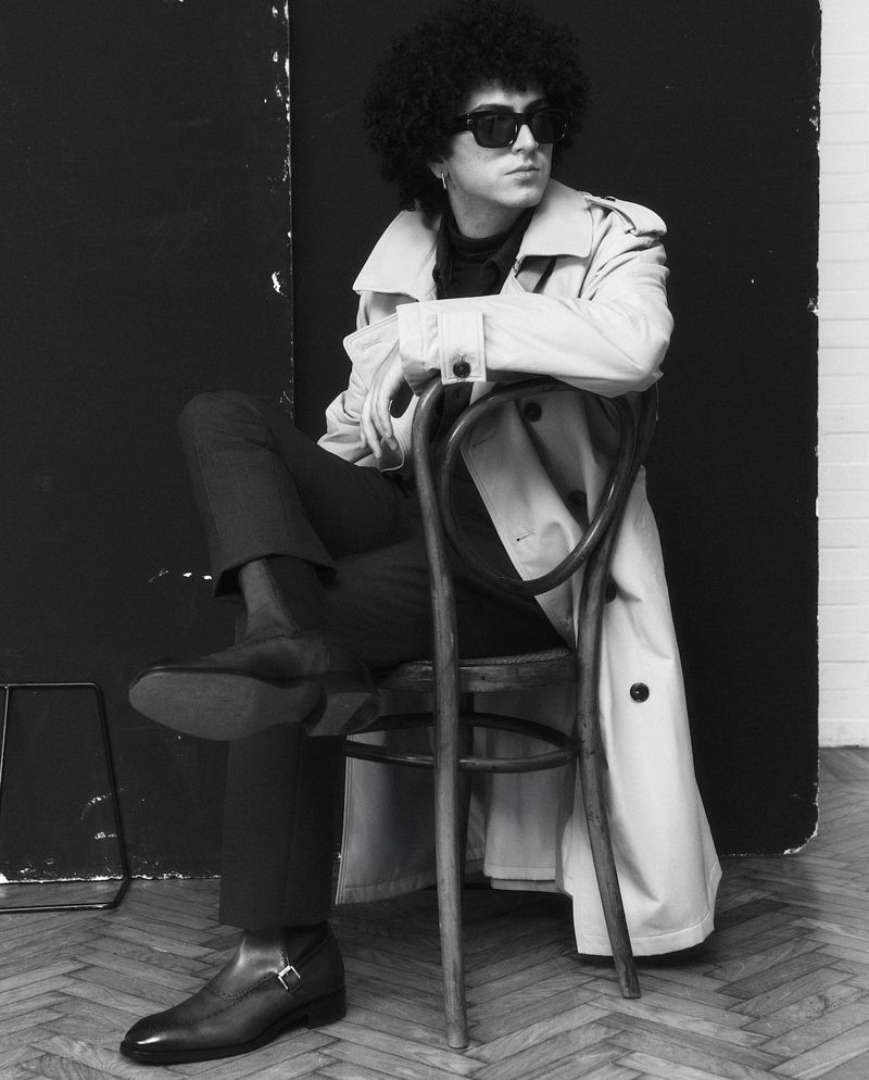 Monochrome portrait of a person with afro hair wearing a trench coat and sunglasses, sitting on a chair in front of a dark backdrop.