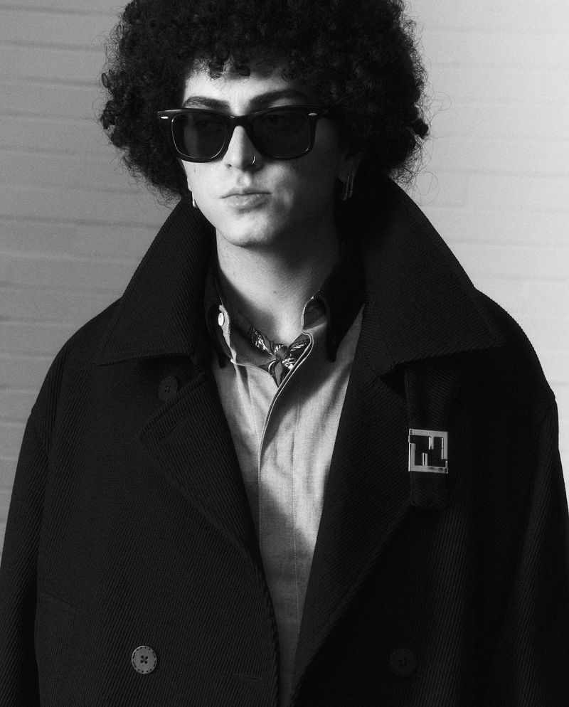 Monochrome portrait of a young adult wearing sunglasses, a light-colored shirt, and a dark overcoat with a logo against a brick wall.